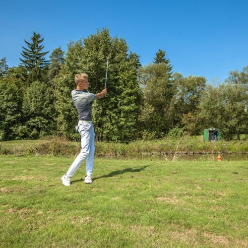 A great shot of a young man playing golf in a field surrounded by trees on a sunny day
