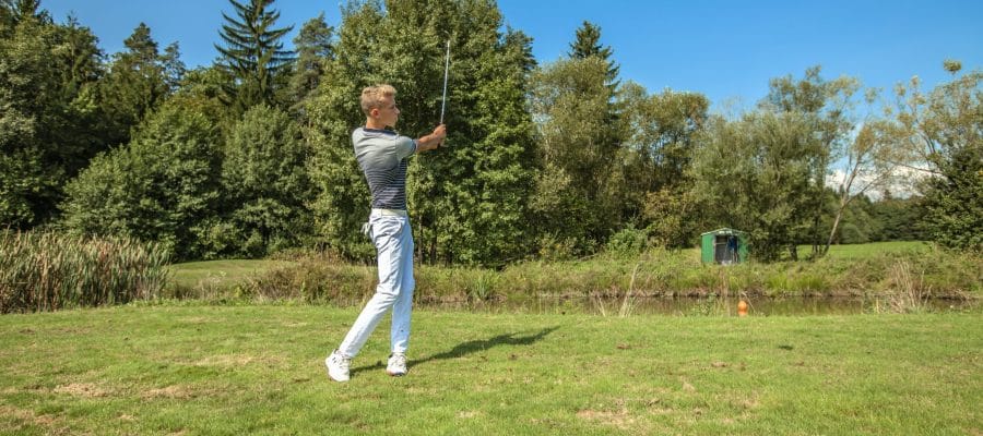 A great shot of a young man playing golf in a field surrounded by trees on a sunny day